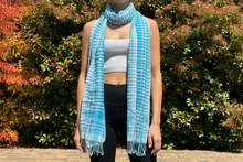 Load image into Gallery viewer, A blue cotton scarf with a checkered pattern, displayed on a mannequin against a backdrop of greenery.
